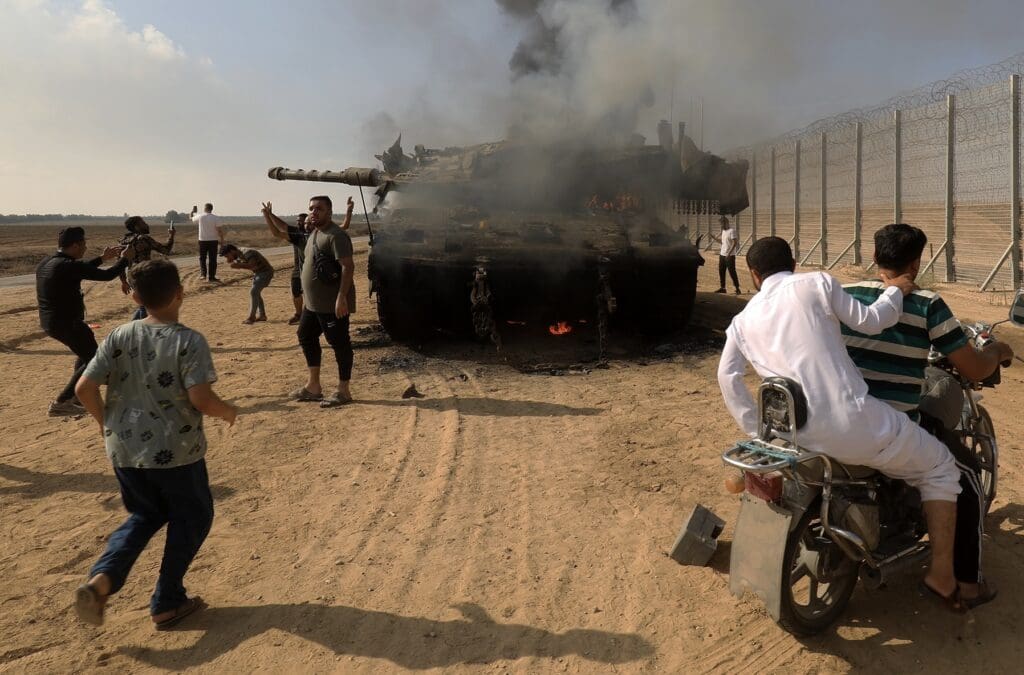 A group of people gather around a burning tank near a fence, some taking photos and others on a motorcycle. The scene is set in a sandy desert area under a partly cloudy sky.