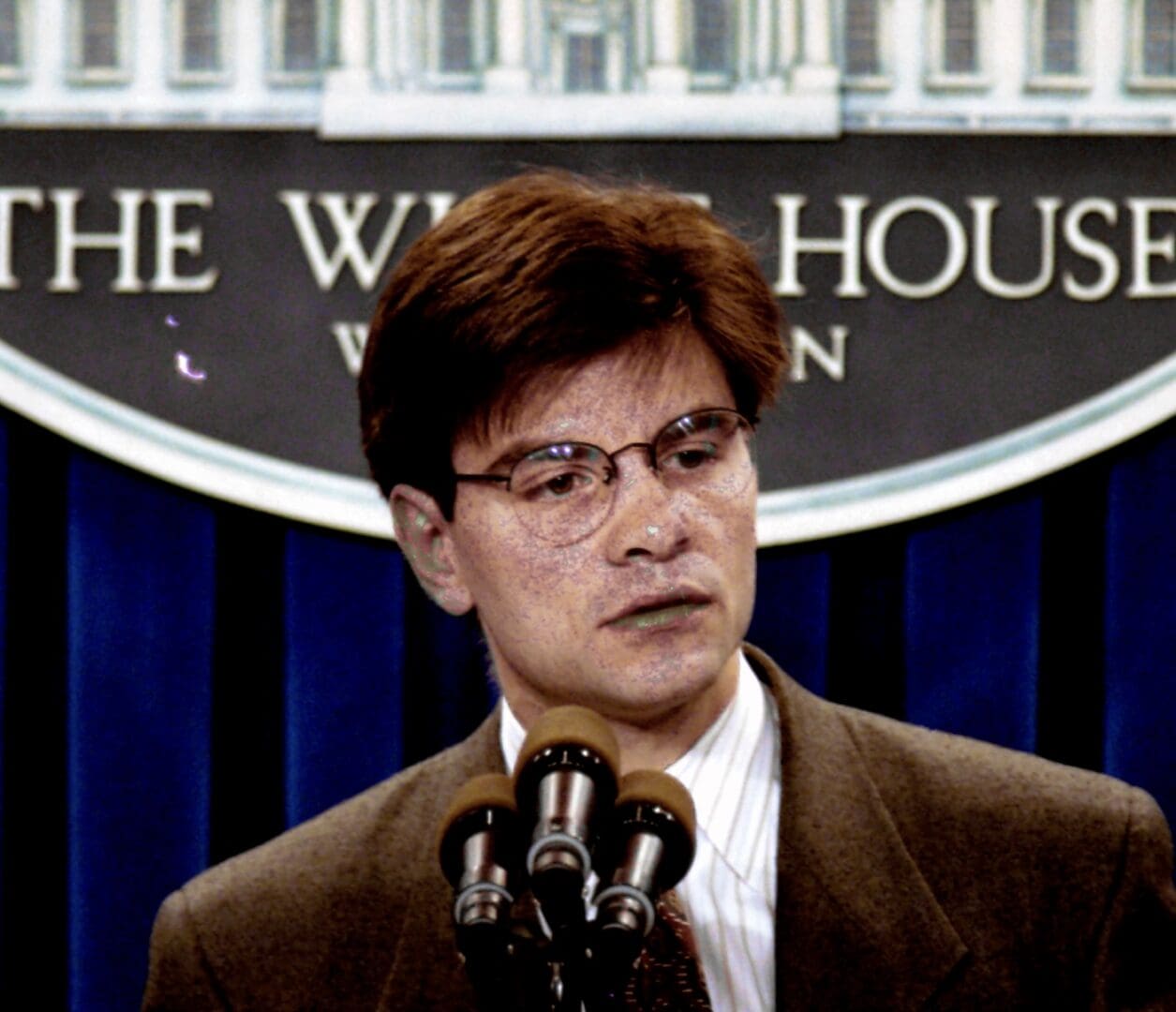 A person in glasses and a suit speaks into multiple microphones